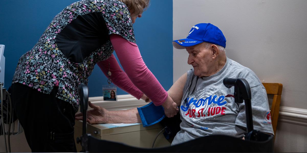 Frank Squibb, one of about 978 people participating in the ARIC study, gets his blood pressure taken by a nurse in colorful scrubs. Frank wears a blue baseball cap and a grey t-shirt.